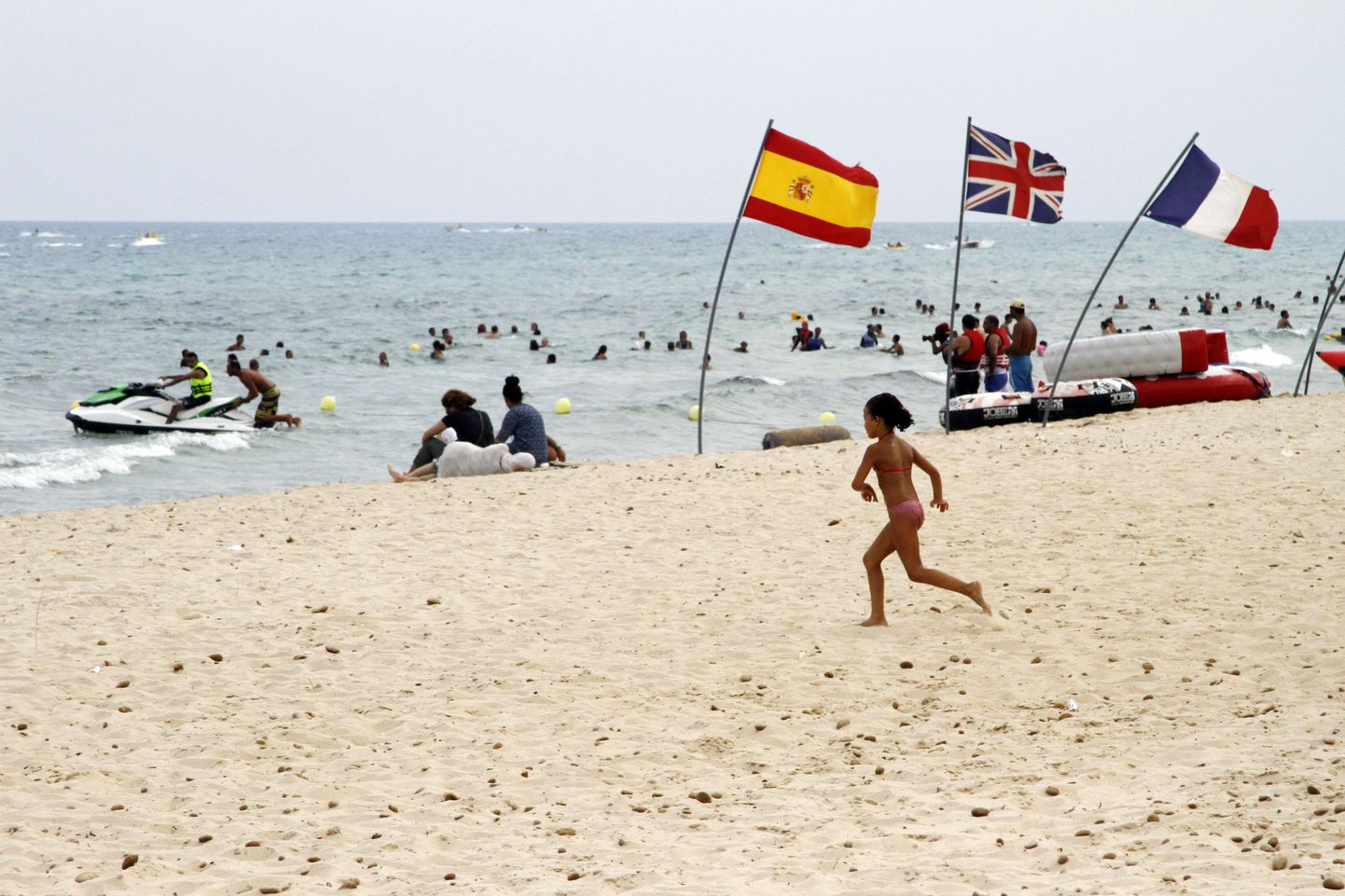 In this Aug. 9, 2015 photo, a young girl runs past European flags on a crowded beach in Hammamet, Tunisia.  The jet skis roar by the shore and the water is full of splashing children along the Tunisian Riviera, where despite the deadly attack that killed 38 tourists in June, the beaches are still full _ for now. Europeans have abandoned this North African country, leaving just local Tunisian beach-goers and visiting Algerians. But they will be gone by months end, and then the real pain for the countrys all-important tourism industry will begin. (AP Photo/Paul Schemm)