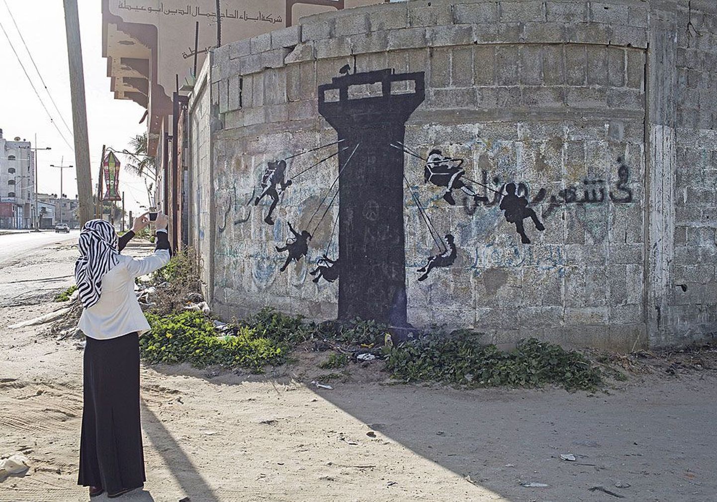 Палестинская женщина снимает картину Бэнкси на главной улице Газа Бейт-Лахийя, на которой 
израильская  сторожевая вышка изображена в виде карусели.