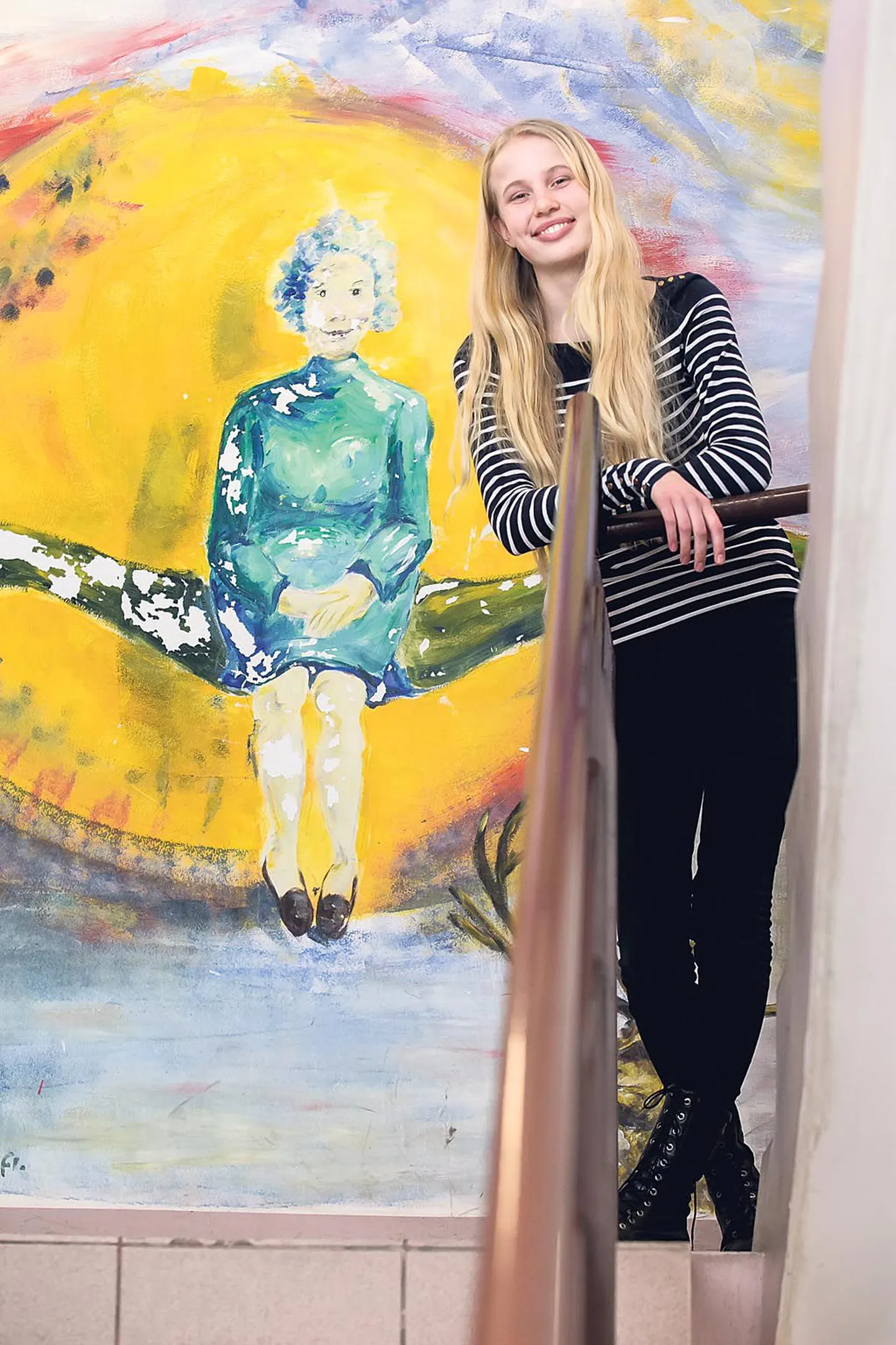 Liisamari Viik poseerib Sütevaka humanitaargümnaasiumis seina taustal, mille on värviliseks maalinud sama kooli vilistlane kunstnik Flo Kasearu.