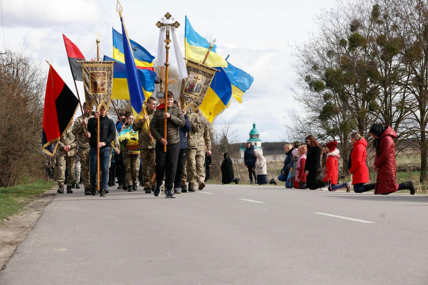Женщины и дети стоят на коленях у дороги, когда мимо них проходит похоронная процессия павшего солдата Владислава Войтовича, направляющаяся в церковь.