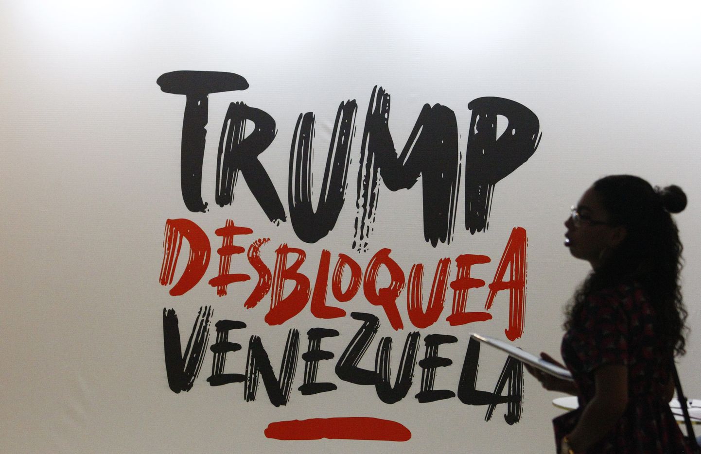Naine kõndimas mööda sildist, mis ütleb hispaania keeles "Trump, võta Venezuela lukust lahti". See viitab Venezuela valitsuse seisukohta, et riigi pikalt kestnud majandusprobleemide taga on USA sanktsioonid, mitte Venezuela võimude küündimatus.