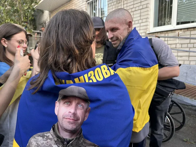 Одна из немногих счастливых пар - Олена (на фото - со спины) с мужем, освободившимся из плена. На фото хорошо видно, как изменился муж Олены за время пребывания в плена. Внизу, на флаге, он запечатлен до плена.