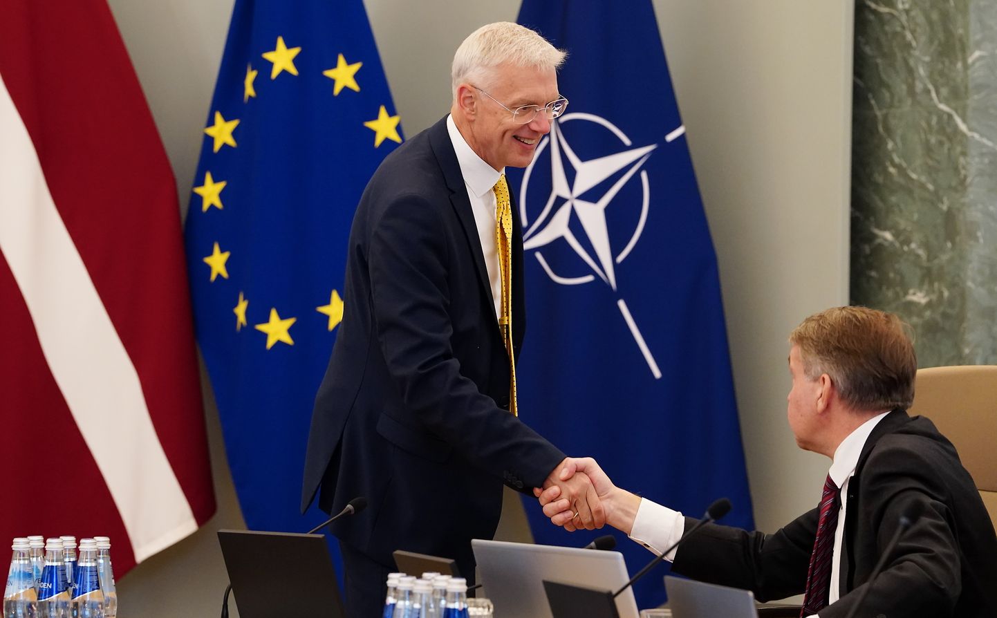 Läti välisminister ja endine peaminister Arturs Krišjānis Kariņš kätleb NATO, ELi ja Läti lipu taustal heaoluministrit Uldis Augulist.