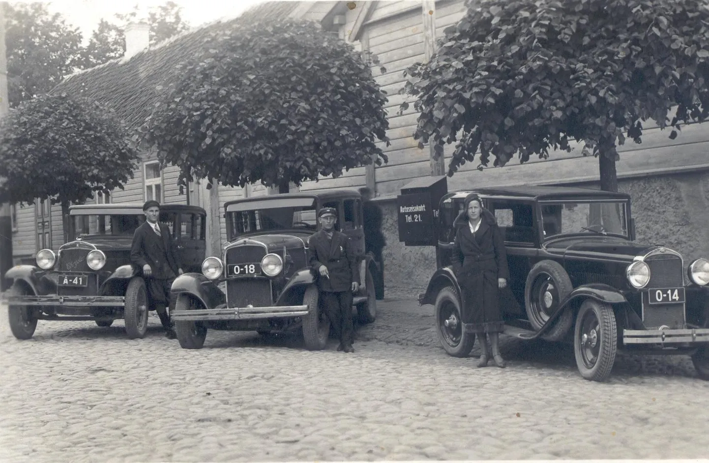 Taksotelefon Võrus. Taksode seisuplatsidele hakati 1930. aastatel paigaldama telefone, kuhu sai taksotellija helistada, et auto endale välja kutsuda. Enamasti said neid telefone raha eest kasutada ka linnakodanikud, kel oli parajasti vaja helistada. Taksojuht pidi telefoni oma kuludega paigaldama ja maksma ka igakuist abonemenditasu. 1928. aastal alustas Tallinnas tööd esimene naistaksojuht Magda Vitismann. Käesolev fotol on teiste kõrval näha ainus Võru naistaksojuht Adele Heuer.